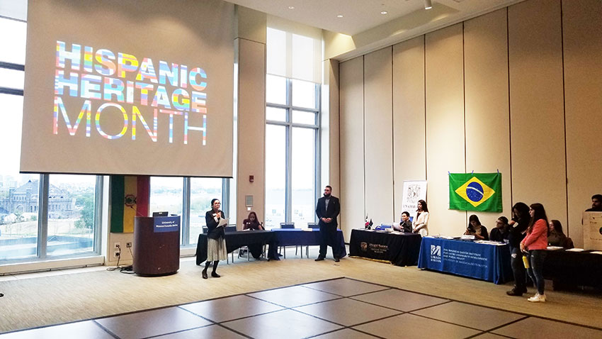 Student speaking in large hall in front of a huge screen that says Hispanic Heritage month 