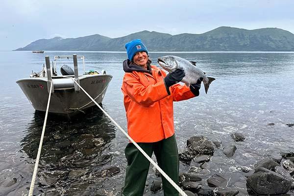 A person i a red coat holding up a big fish while standing next to a boat and a  lake