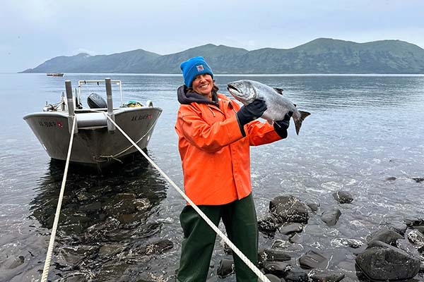 A person i a red coat holding up a big fish while standing next to a boat and a  lake